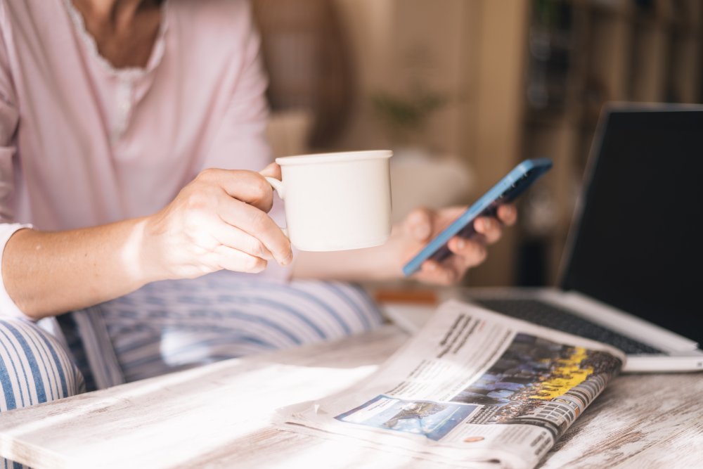 Person med mobil, laptop och morgontidning. 