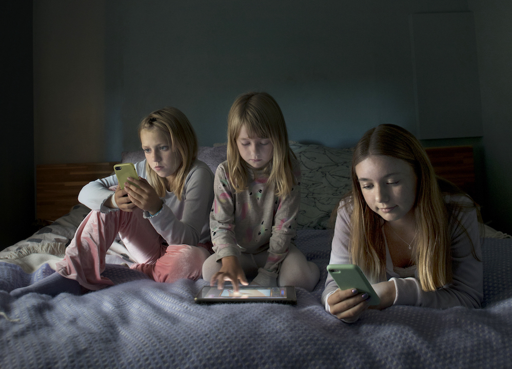Three kids sitting on a bed using Ipads and smartphones.