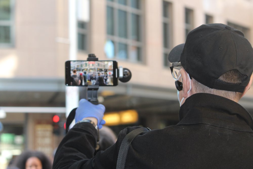 Journalist filmar en grupp människor, samtidigt som han bär mask. 