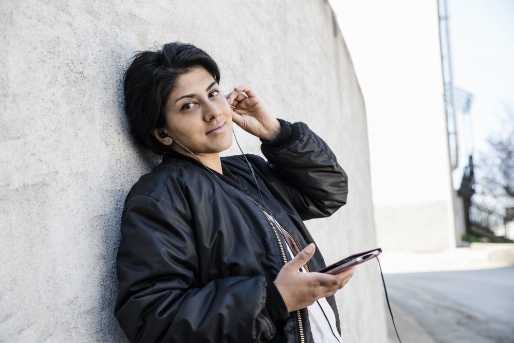 Young woman leaning agains a wall listening to her mobile phone via earplugs